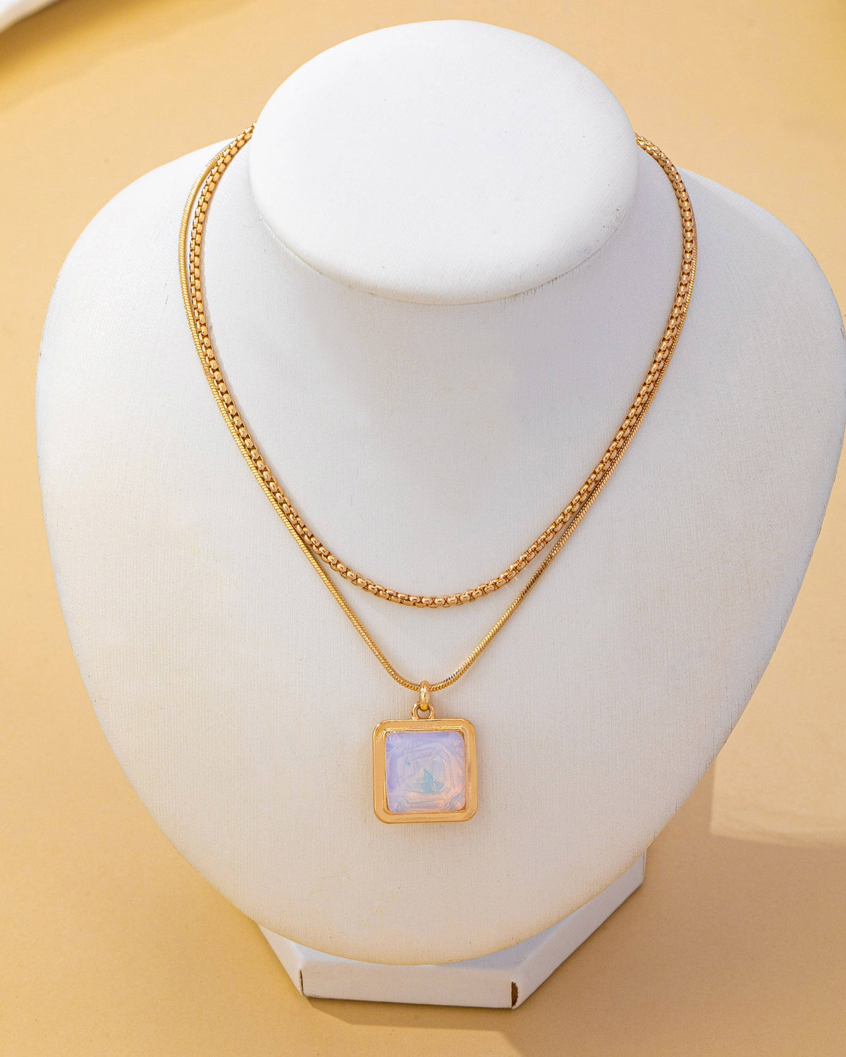 A gold double-layer necklace with a square iridescent opal pendant, displayed on a white jewelry bust against a warm beige background.