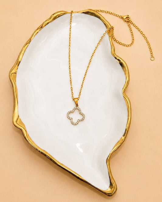 Gold clover and crystal open pendant on gold chain in a gold lined white jewelry bowl on beige background.