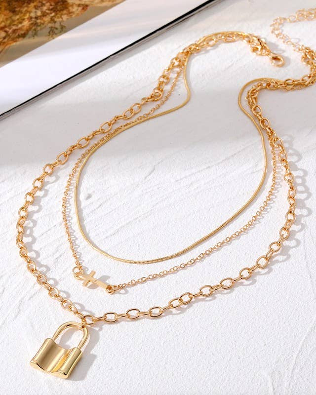 Gold layered chain necklace with a padlock pendant and a cross charm on a white surface.