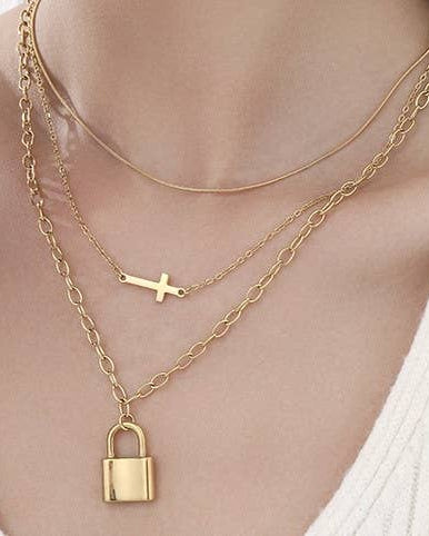 A close-up of a person's neck and upper chest wearing a layered gold necklace set. The set includes a delicate chain, a chain with a small cross pendant, and a longer chain with a padlock pendant.