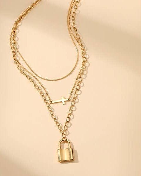 A gold-toned layered necklace featuring three chains, a small cross charm, and a padlock pendant, displayed on a warm beige background.