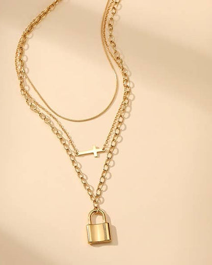A gold-toned layered necklace featuring three chains, a small cross charm, and a padlock pendant, displayed on a warm beige background.