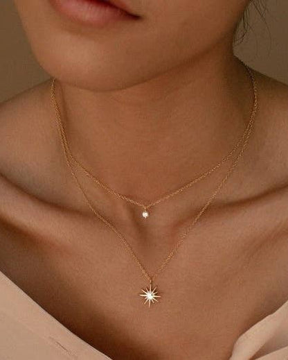A close-up of a model wearing a delicate double-layered necklace. The necklace features a fine gold chain with a small starburst pendant and a second layer with a small CZ. 