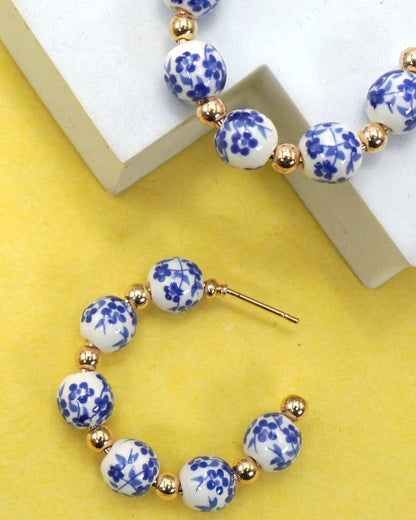 Hoop earrings with blue floral toile porcelain beads and gold accents on a yellow background.
