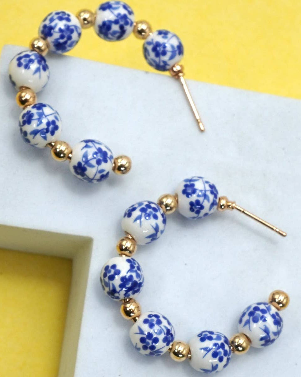 A pair of beaded hoop earrings with white ceramic beads featuring blue floral patterns and small gold spacer beads, displayed on a white geometric stand against a yellow background.