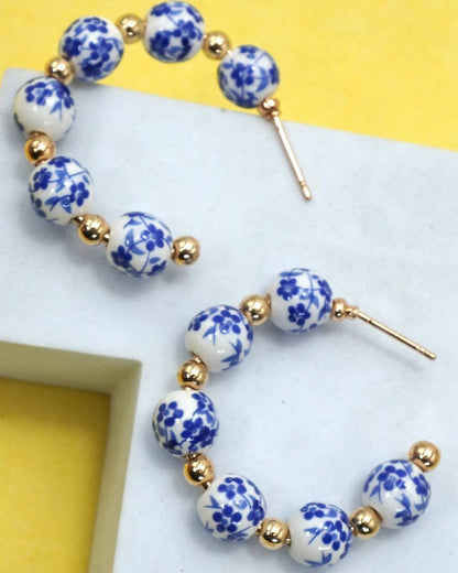 A pair of beaded hoop earrings with white ceramic beads featuring blue floral patterns and small gold spacer beads, displayed on a white geometric stand against a yellow background.