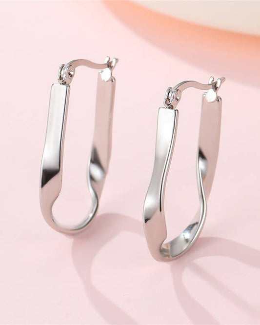 A pair of polished silver hoop earrings with a modern, wavy open design, displayed on a soft pink background.