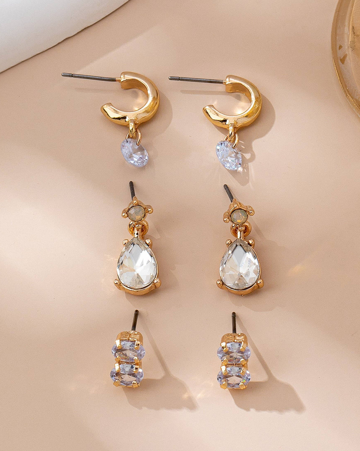Trio of gold earrings with clear rhinestones on a beige surface