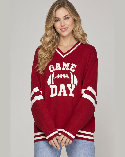 A woman modeling a red V-neck knit sweater with 'GAME DAY' and a football graphic in white on the front, featuring white sleeve and hem stripes, paired with light blue jeans against a light gray studio background.