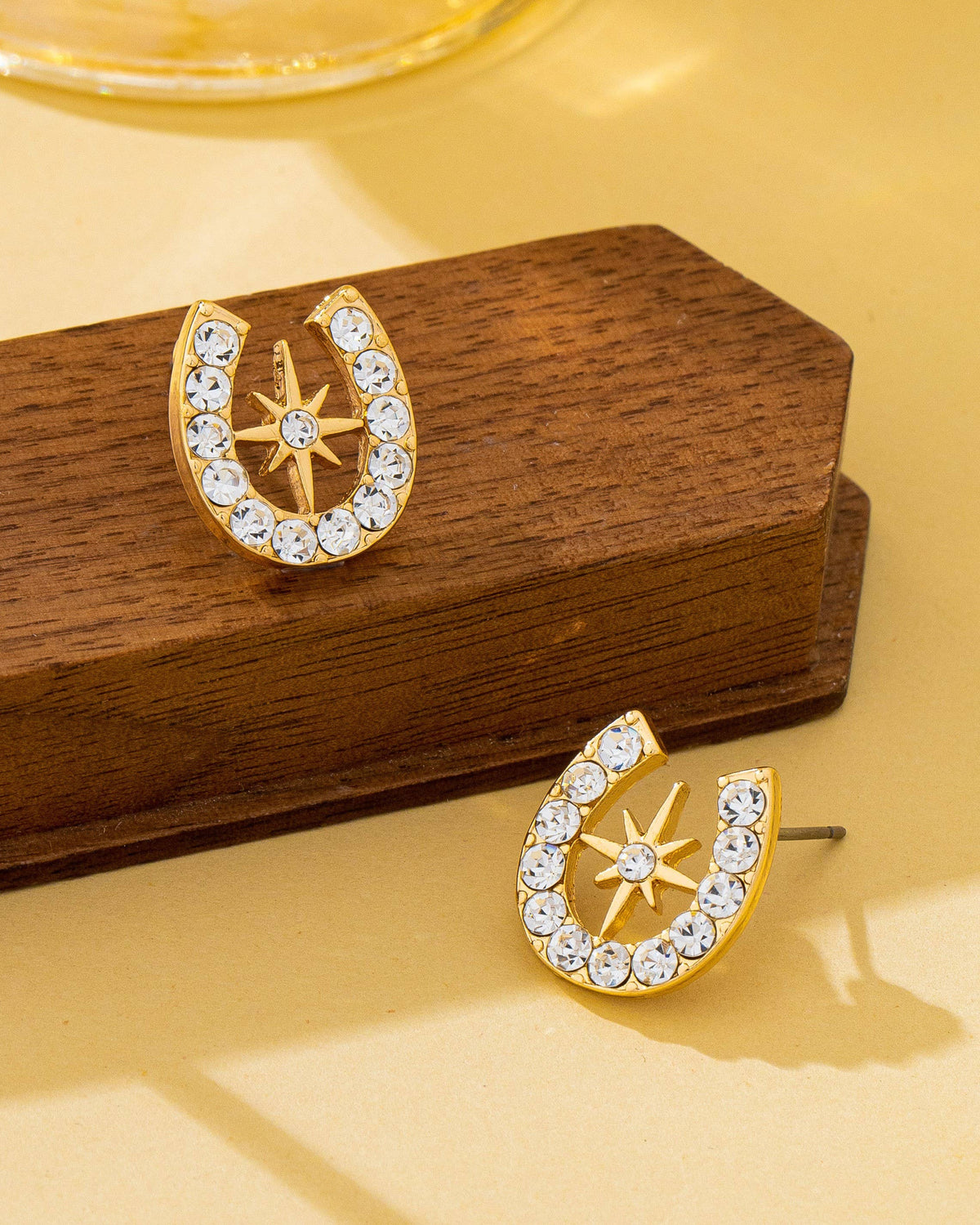 Gold earrings with star and horseshoe design on a wooden stand against a yellow background.