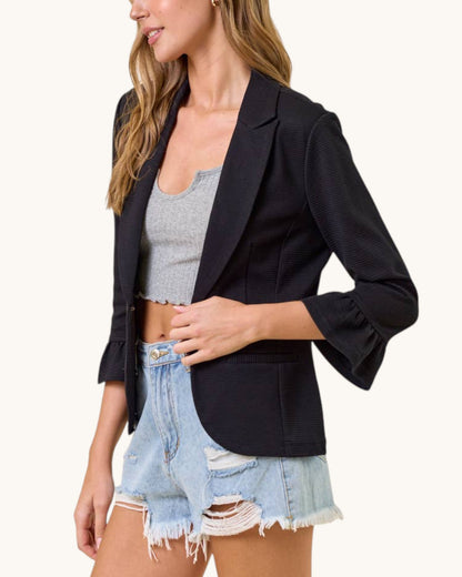 A woman wearing a black textured blazer with flared three-quarter sleeves, styled with a gray crop top and light-wash distressed denim shorts, against a solid off-white background.