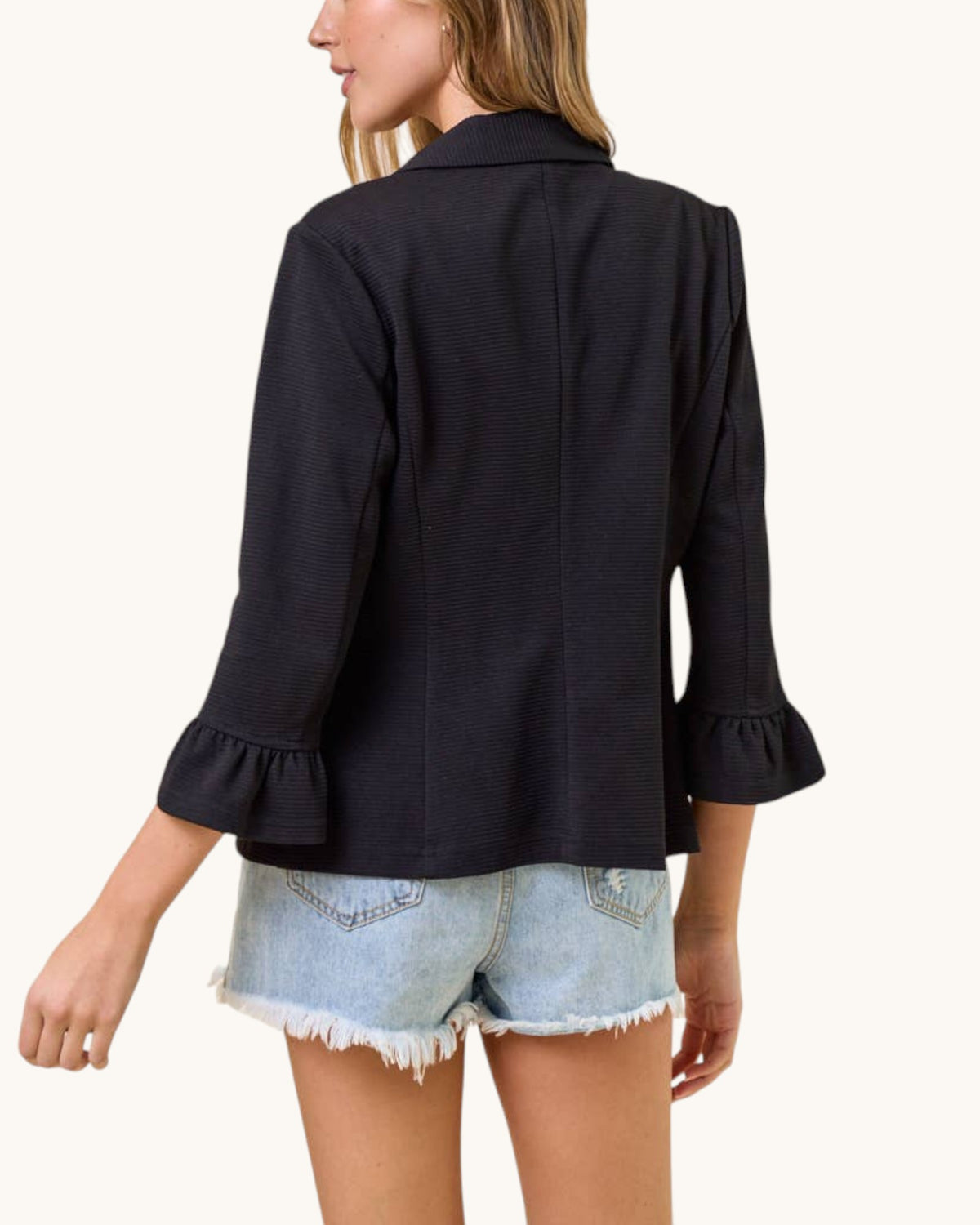 A woman shown from the back wearing a black textured three-quarter sleeve blazer with ruffled cuffs and light-wash denim shorts against a plain off-white background.