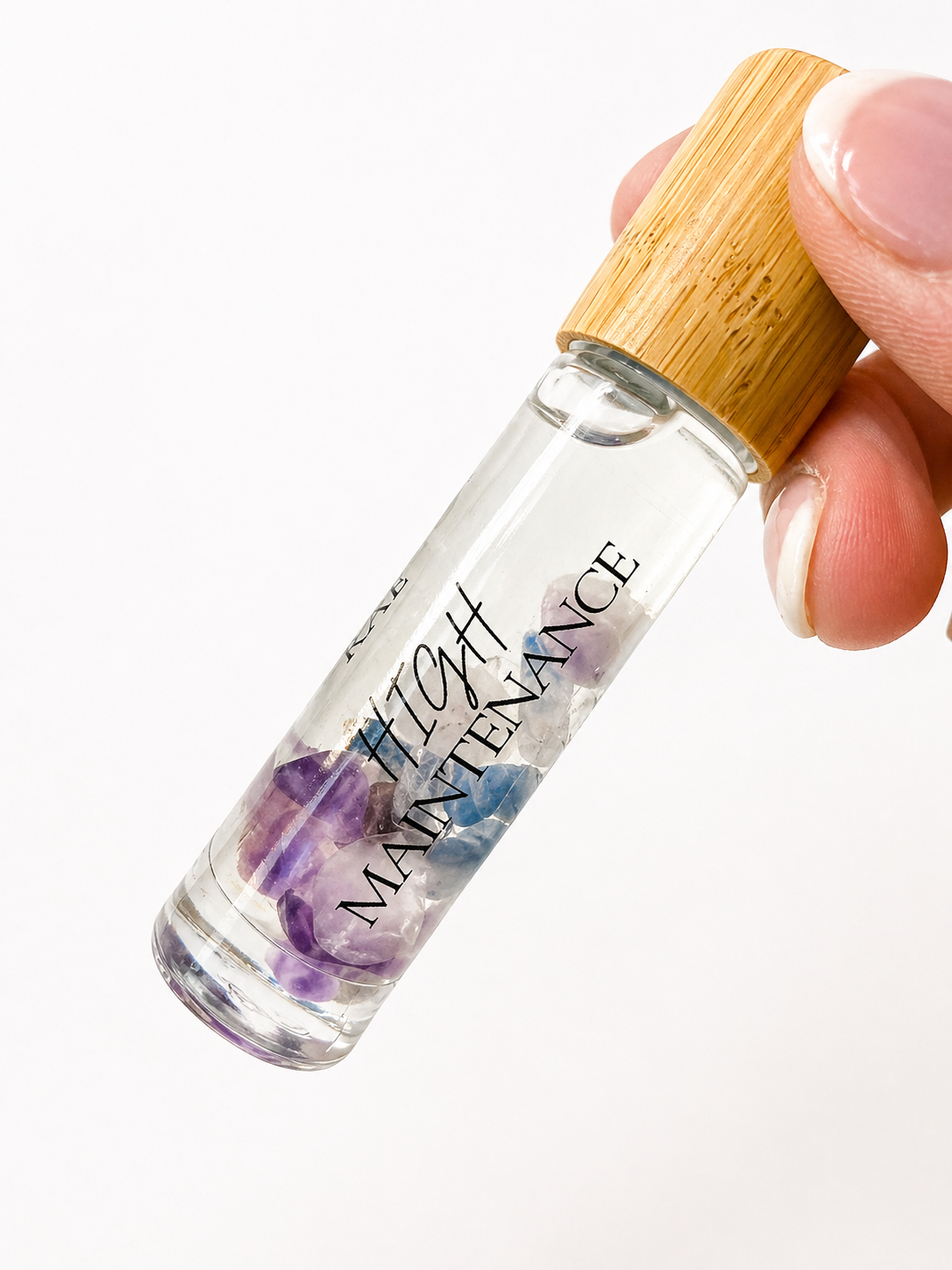 Small clear bottle with bamboo cap labeled 'High Maintenance' held by a hand on a white background.
