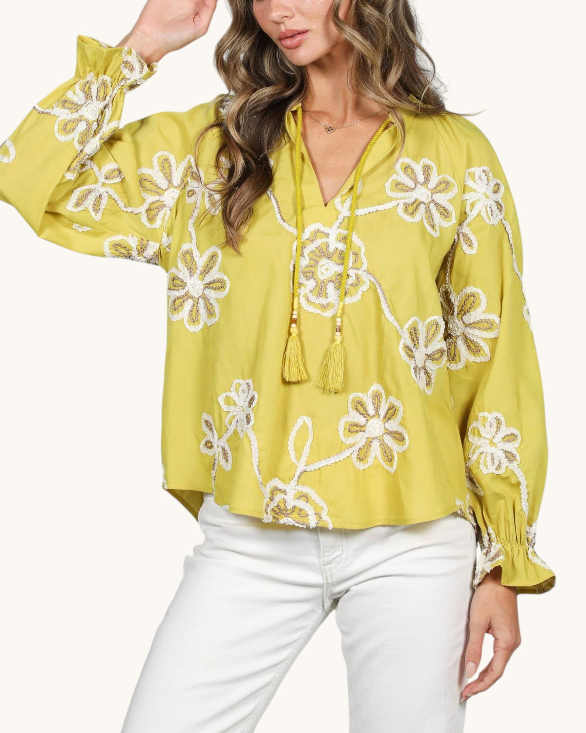 A woman wearing a greenish-yellow long-sleeve blouse with white floral 3-D embroidery and tassel ties, paired with white pants, against a light beige background.
