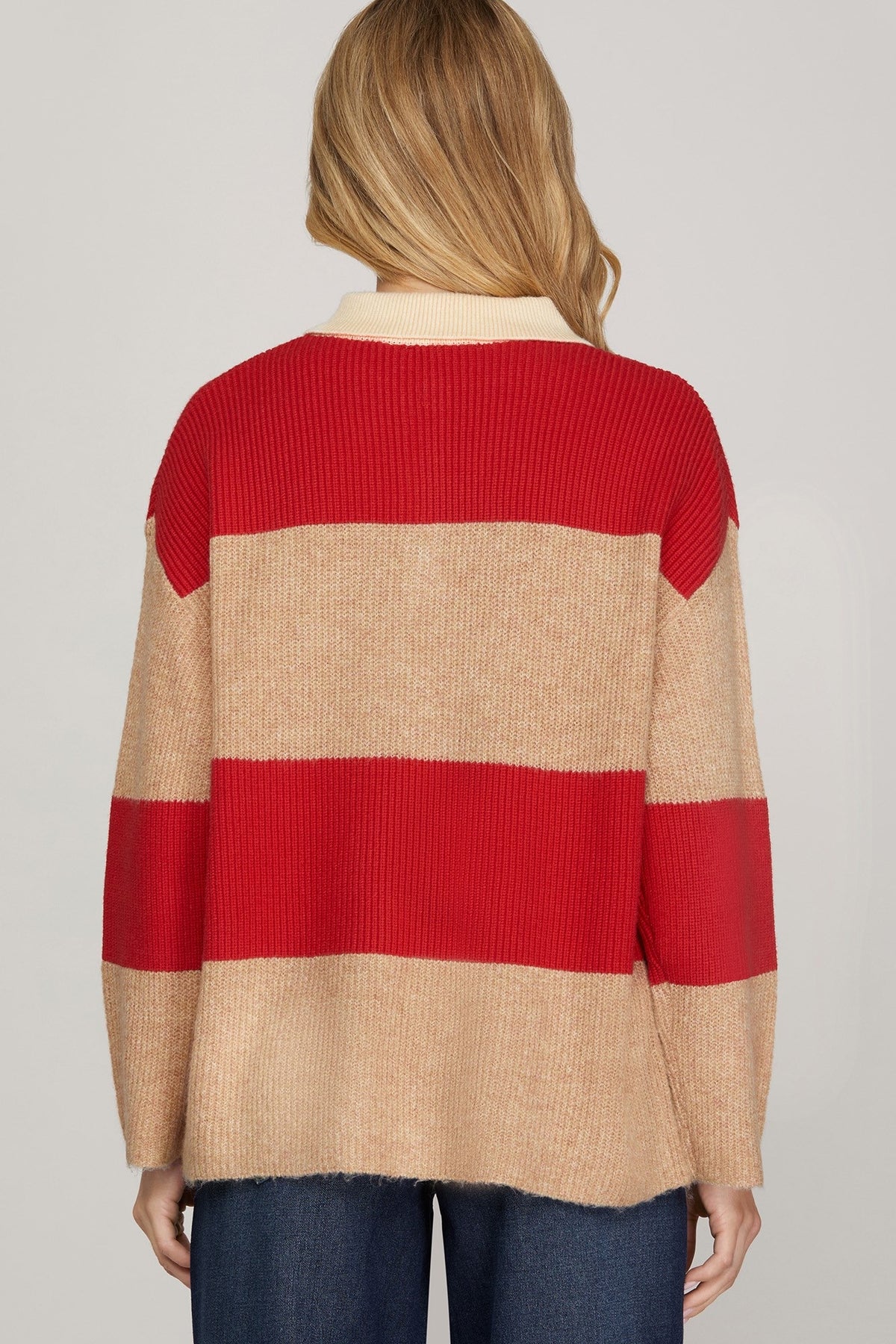 A woman viewed from the back wearing a ribbed knit sweater with wide horizontal stripes in red and beige, paired with dark blue jeans, against a light gray studio background.