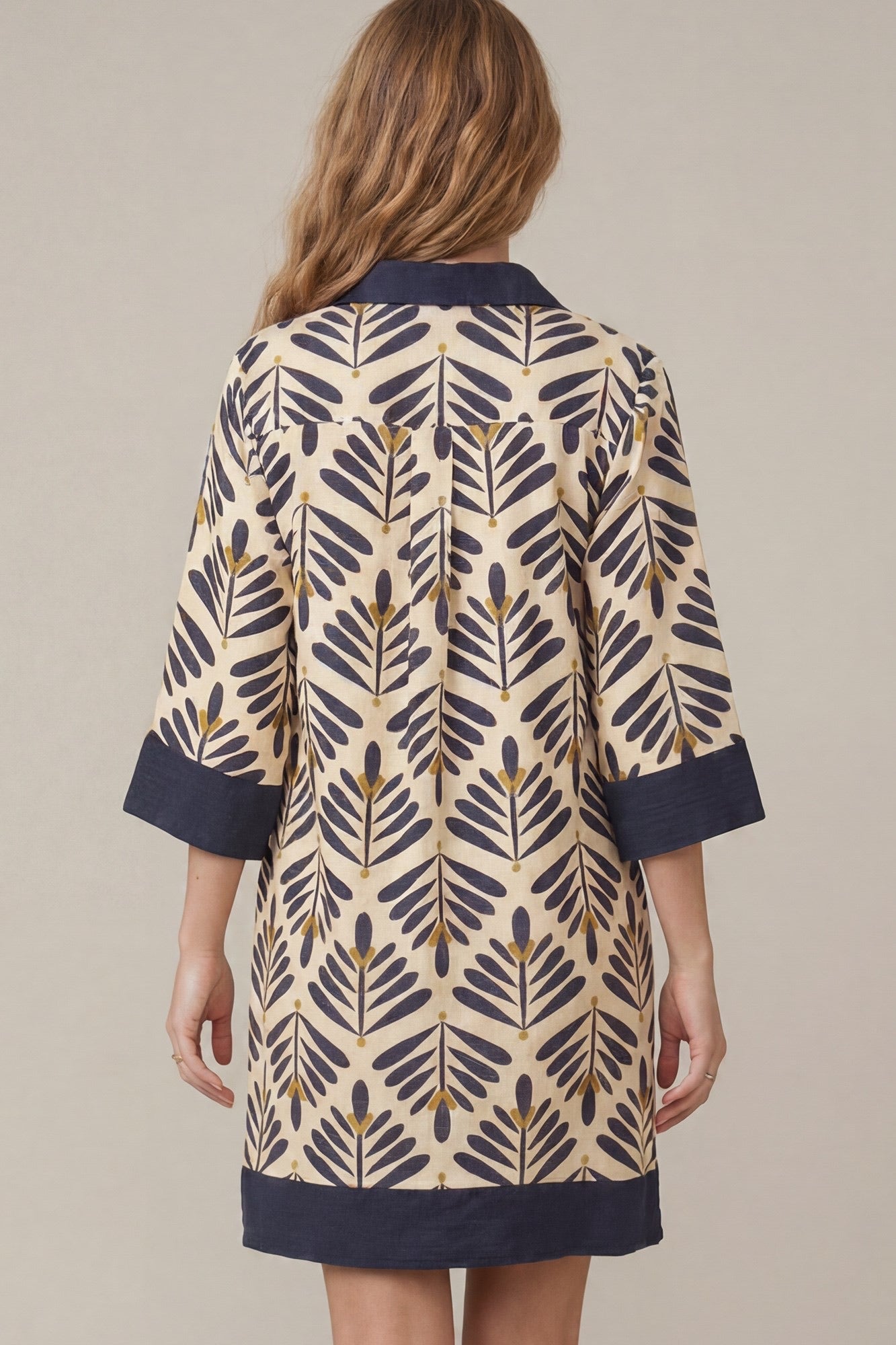 A woman viewed from the back wearing a short tunic or dress with a cream base and an allover dark navy leaf pattern, finished with solid navy trim at the collar, cuffs, and hem.