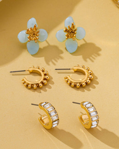 Three pairs of gold-tone stud and hoop earrings arranged on a warm beige surface. The top pair features light green flower-shaped stones with gold centers, while the other pairs are gold hoops, one with small bead detailing and one with clear rhinestones.