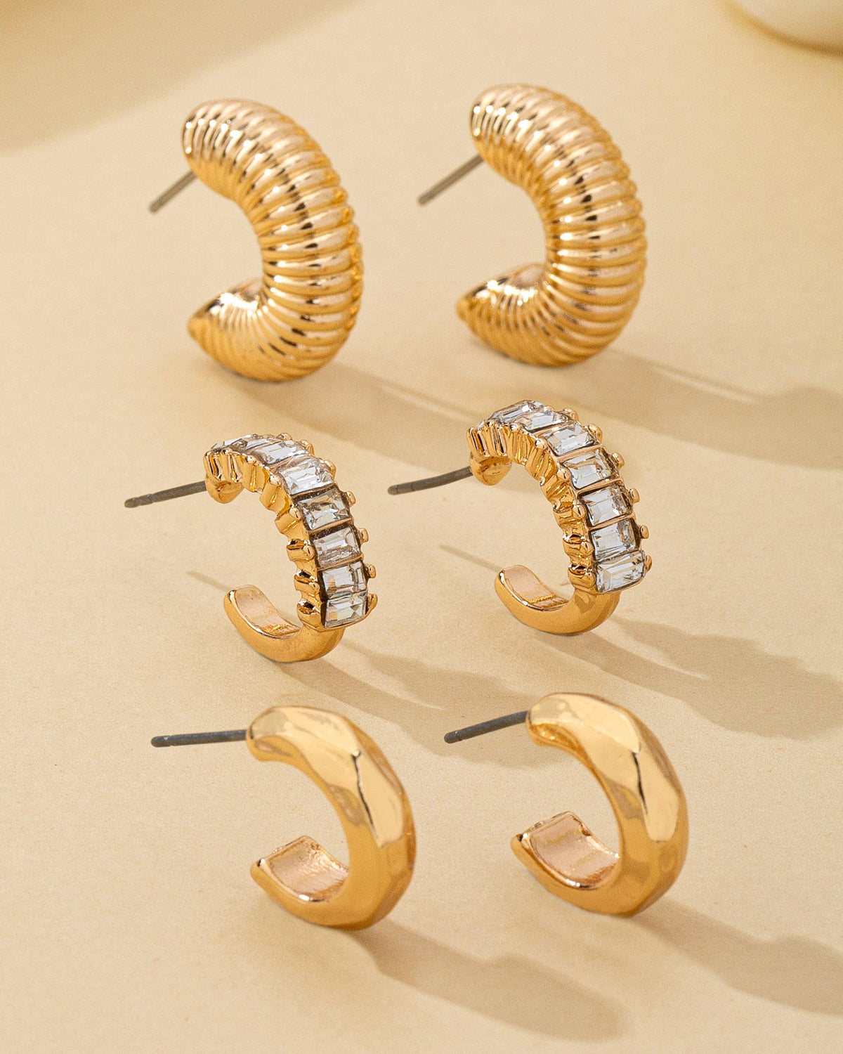 Trio of gold hoop earrings on a beige background with crystal accents and textured finishes.