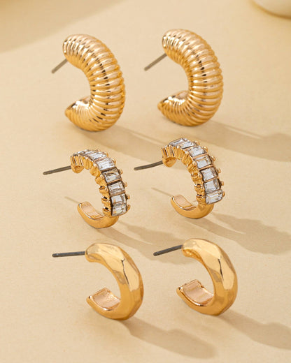 Trio of gold hoop earrings on a beige background with crystal accents and textured finishes.