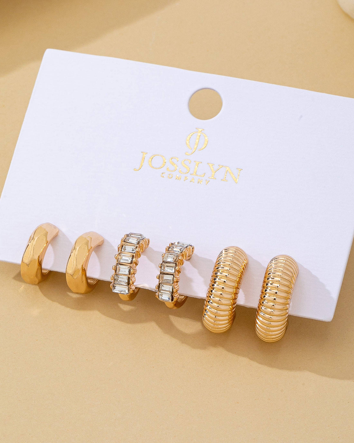 Set of gold earrings featuring 3 textured and crystal hoops on a Josslyn card against a beige background.