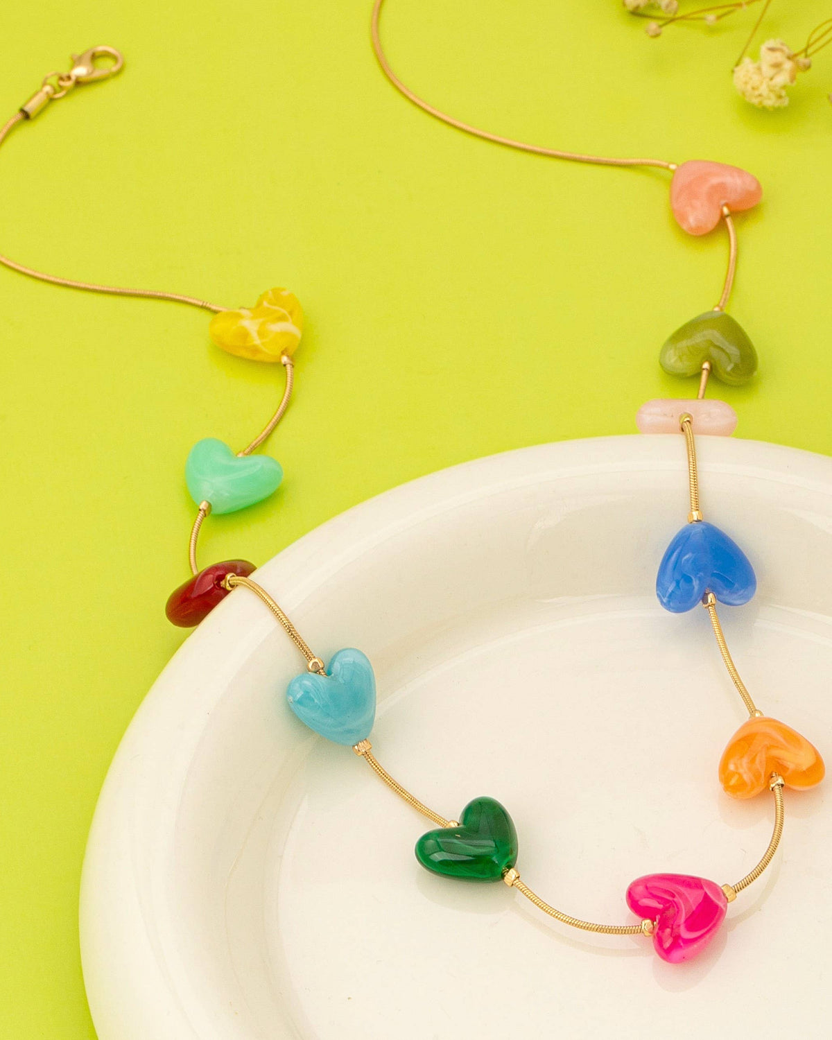 A gold-tone necklace with multiple translucent heart-shaped beads in assorted bright colors, displayed on a white plate against a bright yellow-green background.