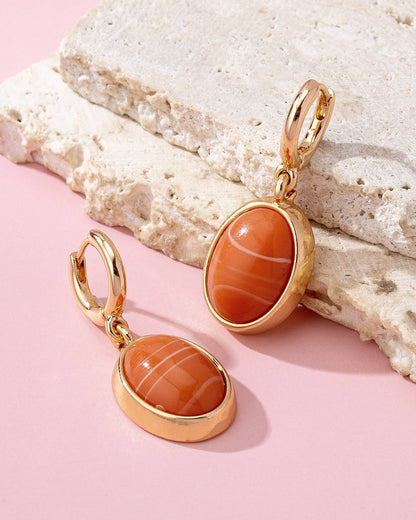 Gold earrings with orange stones on a textured stone surface with a pink background.