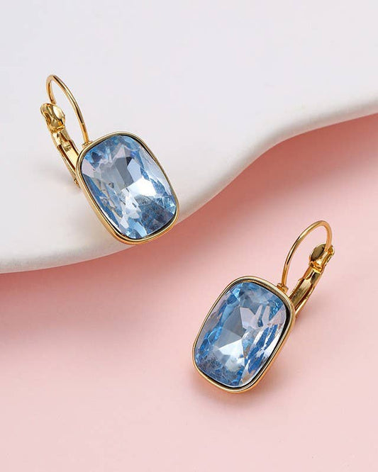 A pair of gold leverback earrings with rectangular light blue faceted gemstones/crystals, displayed on a wavy white and pink background.