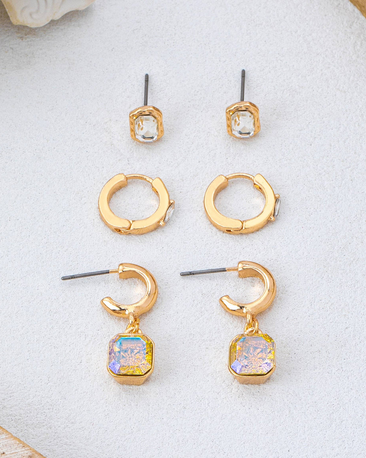 Three pairs of gold-toned crystal earrings arranged on a light textured surface: small square stud earrings with clear stones, huggie hoop earrings, and C-shaped drop earrings with iridescent square stones.