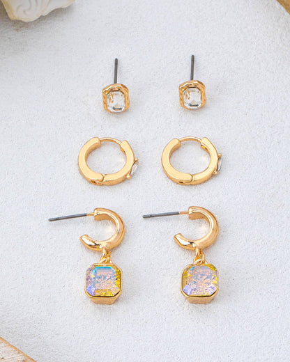 Three pairs of gold-toned crystal earrings arranged on a light textured surface: small square stud earrings with clear stones, huggie hoop earrings, and C-shaped drop earrings with iridescent square stones.