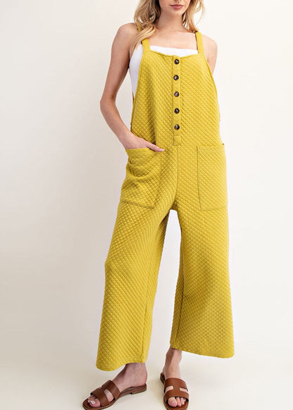 A woman modeling a yellow/green sleeveless jumpsuit with front pockets and a button-up front, styled with a white tank top underneath and brown slide sandals against a plain light beige studio background.