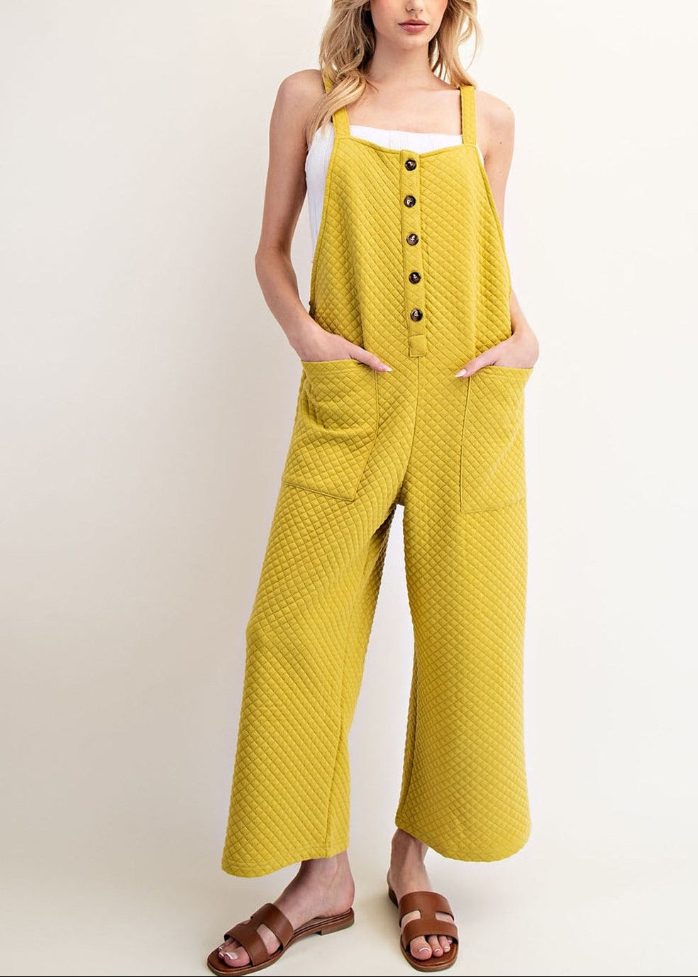A woman modeling an avocado yellow wide-leg jumpsuit with a textured pattern, front button closure, and side pockets, paired with a white tank top and brown sandals against a plain beige background.