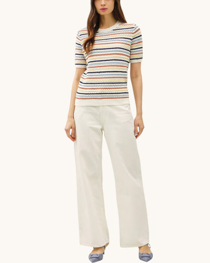 A model wearing a short-sleeve knit top with horizontal multicolor stripes (blue, orange, yellow, and white) paired with off-white wide-leg trousers and blue shoes, set against a solid off-white background.