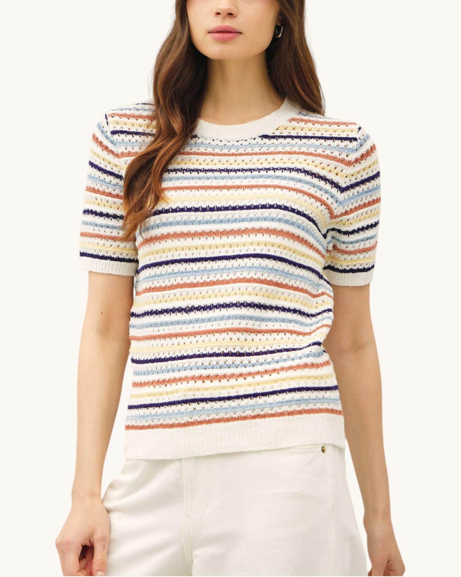 A woman wearing a short-sleeve knit sweater with horizontal multicolor stripes in cream, navy, orange, and light blue, paired with white pants, against a plain off-white background.