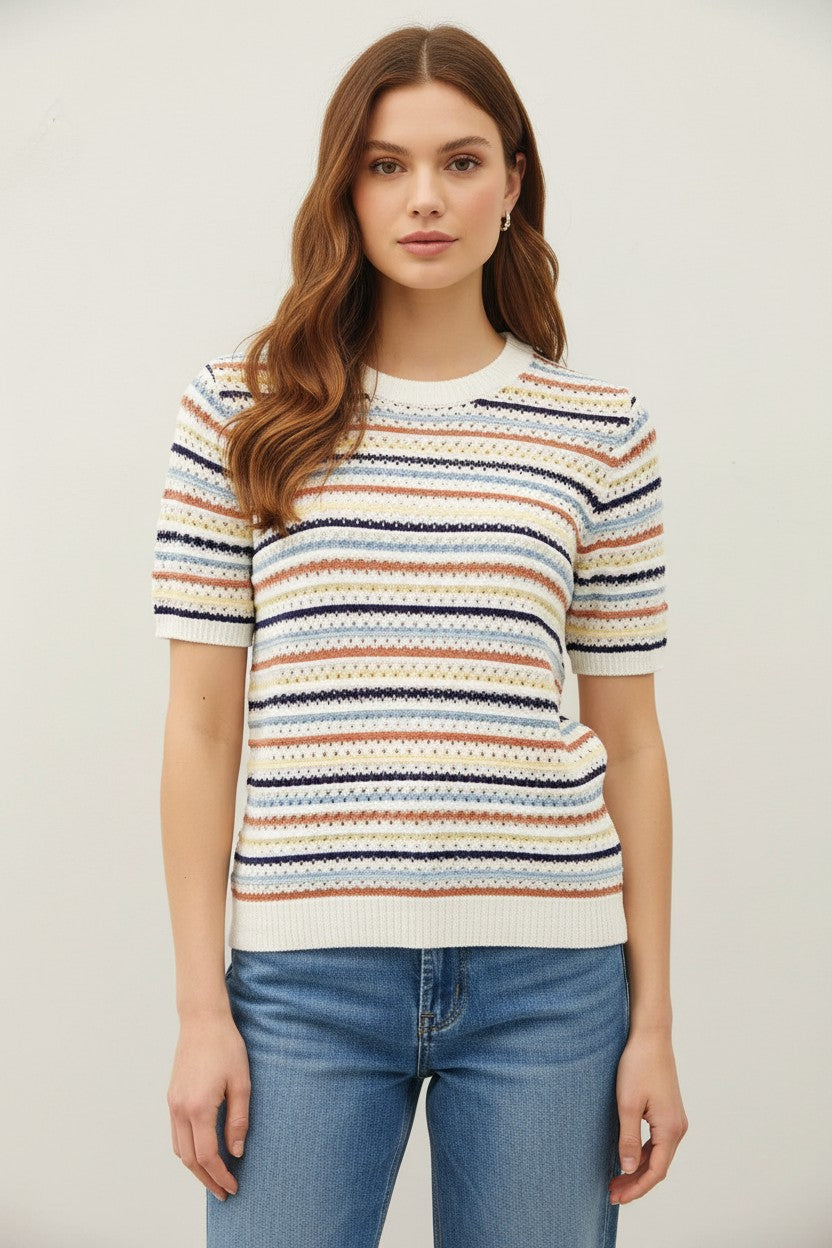 A woman wearing a short-sleeve knit sweater with horizontal multicolor stripes in navy, orange, beige, and white, paired with blue jeans, against a plain light gray background.