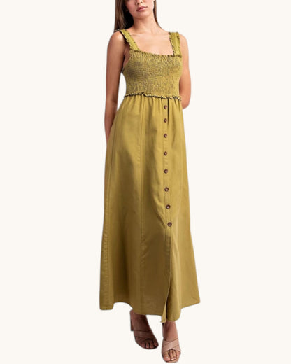 A woman modeling a sleeveless olive green maxi dress with a smocked bodice, ruffled straps, and a button-down front, set against a plain off-white background.