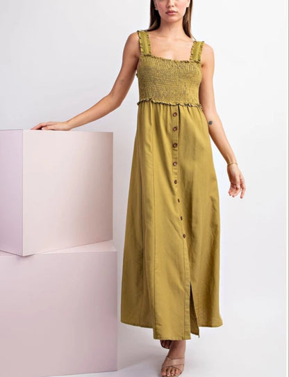 A woman models a olive green sleeveless maxi dress with a smocked bodice, ruffled trim, and a button-down front. She is standing next to a light pink display block against a white studio background.