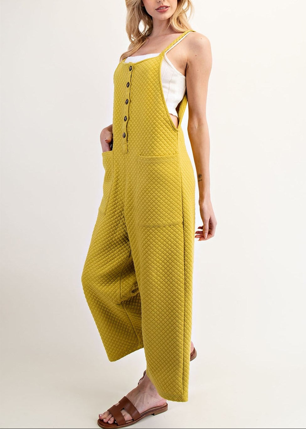 A woman modeling a textured avocado yellow sleeveless jumpsuit with wide legs, front buttons, and side pockets, paired with a white undershirt and brown sandals, against a plain light beige studio background.