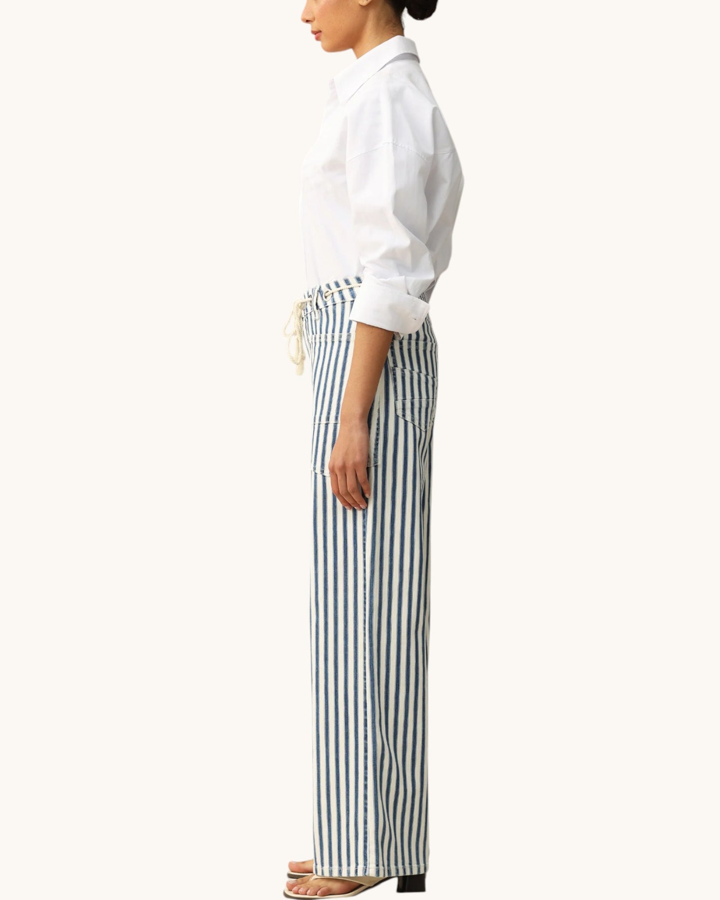 A side-profile view of a model wearing high-waisted, wide-leg trousers with vertical navy and white stripes, paired with a white button-down shirt and flat sandals, against a solid off-white background.