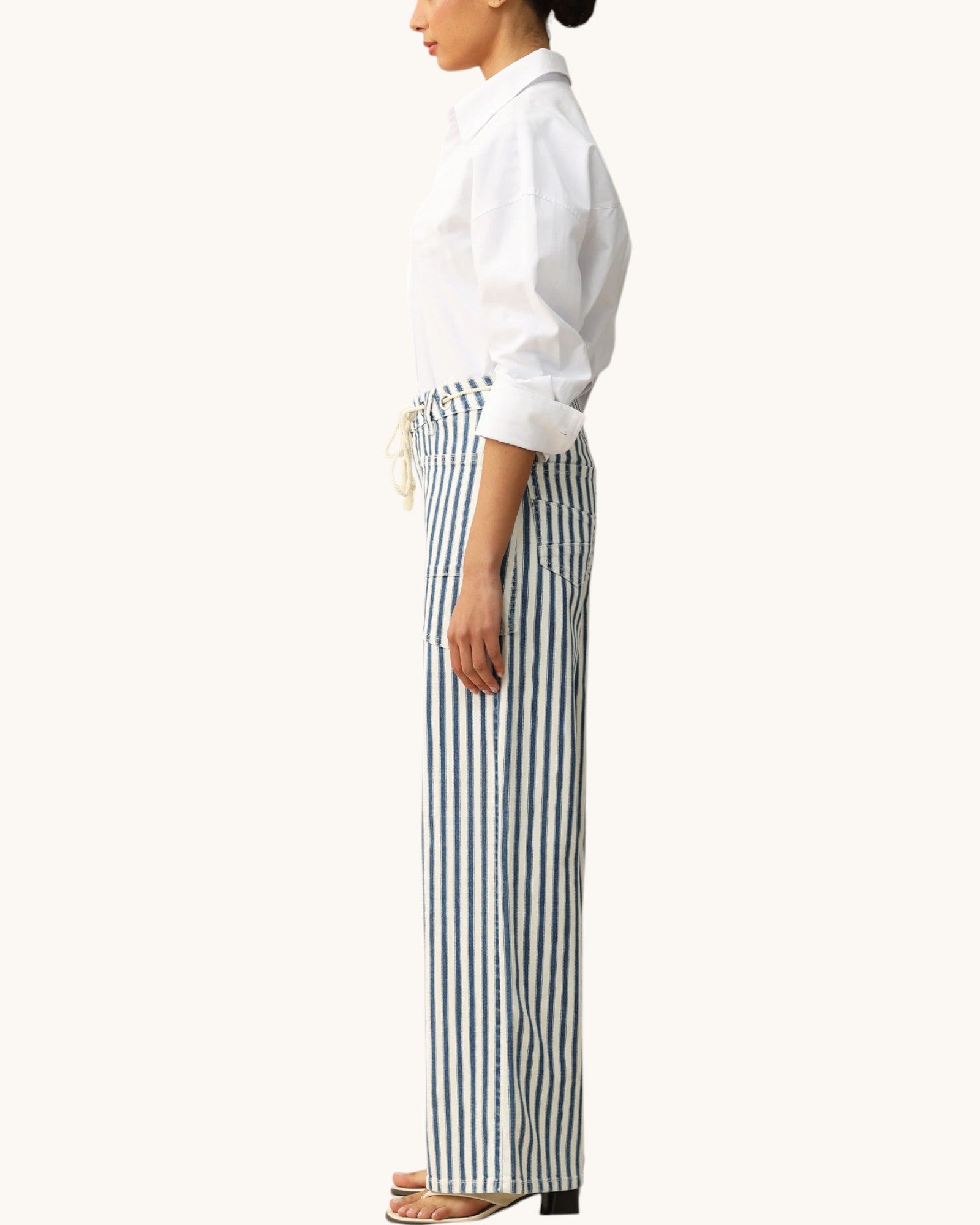 A side-profile view of a model wearing high-waisted, wide-leg trousers with vertical navy and white stripes, paired with a white button-down shirt and flat sandals, against a solid off-white background.