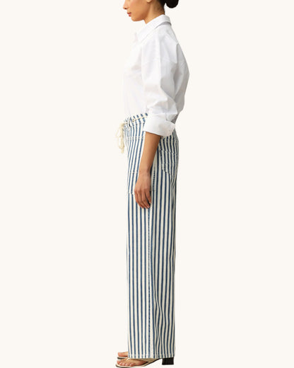 A side-profile view of a model wearing high-waisted, wide-leg trousers with vertical navy and white stripes, paired with a white button-down shirt and flat sandals, against a solid off-white background.