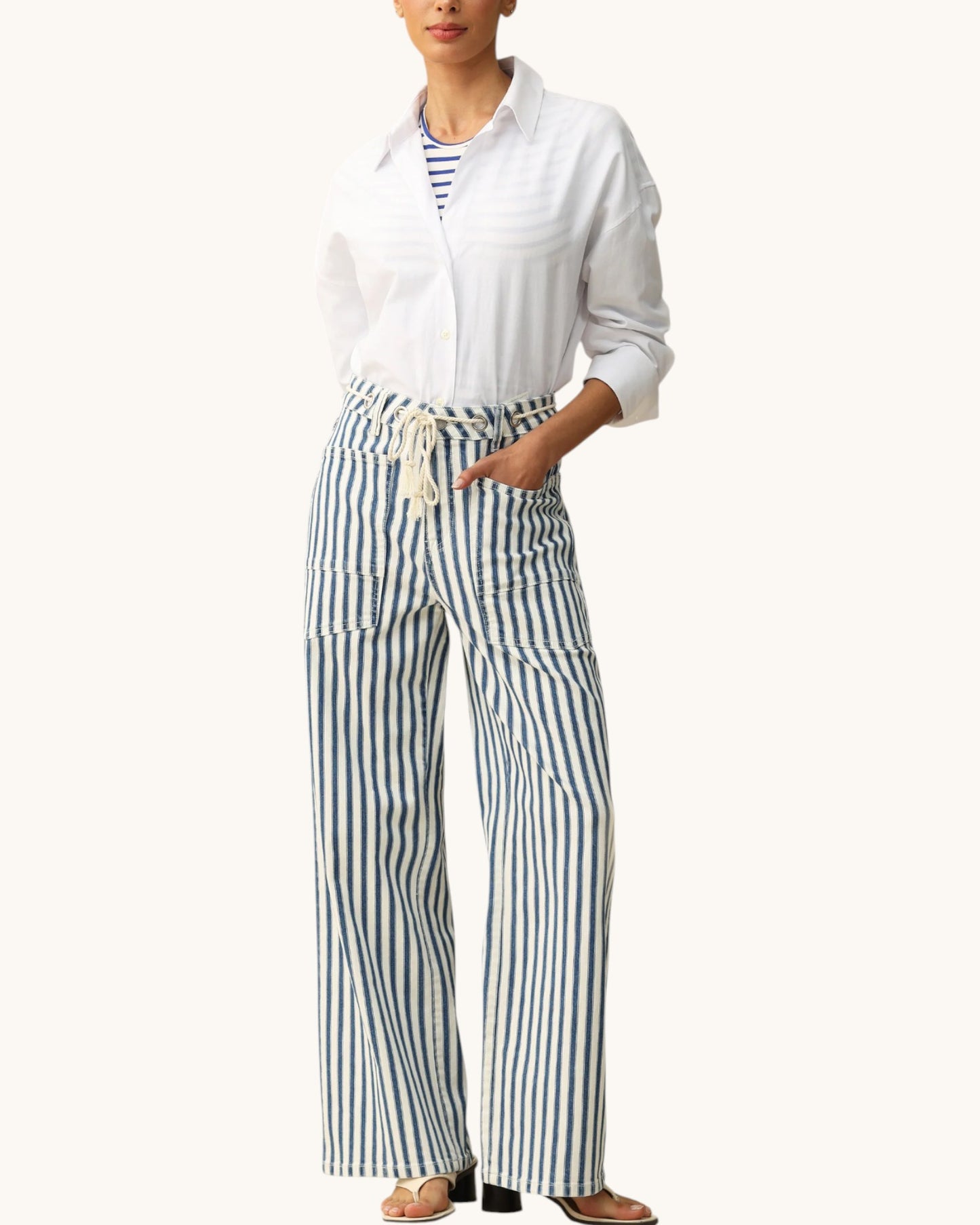 A model wearing a white long-sleeve button-up shirt and high-waisted, wide-leg trousers with vertical blue and white stripes, styled with a matching striped top underneath. The photo is taken against a solid off-white background.