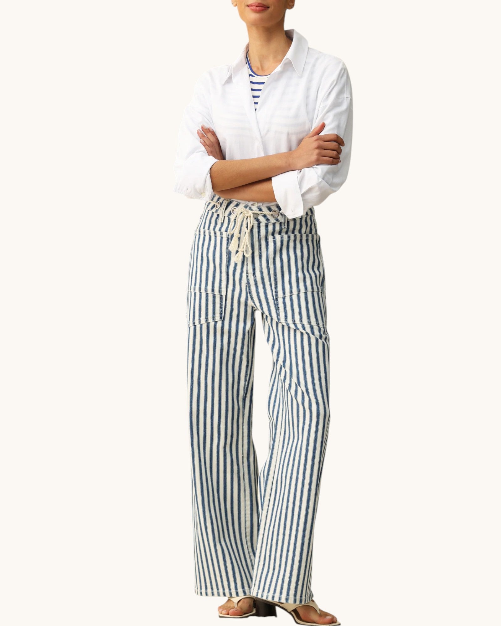 A model wearing a white long-sleeve button-up shirt and blue and white vertically striped wide-leg pants with a drawstring waist, paired with brown sandals, against a solid off-white background.