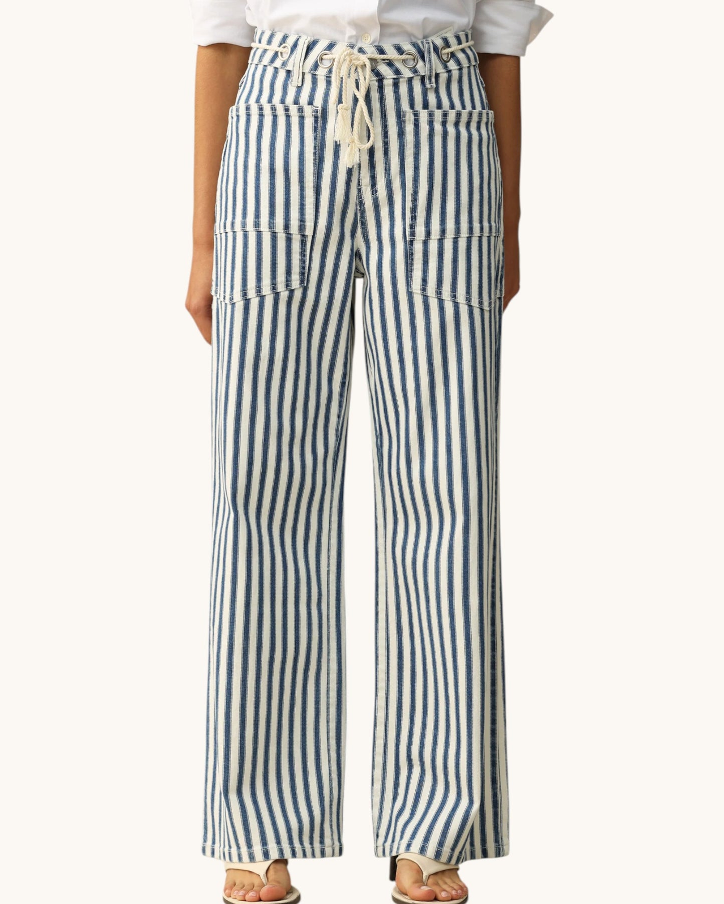 A person wearing high-waisted, wide-leg pants with vertical blue and white stripes, a cream drawstring waist, and large front patch pockets, paired with a white top and tan sandals against a plain light background.