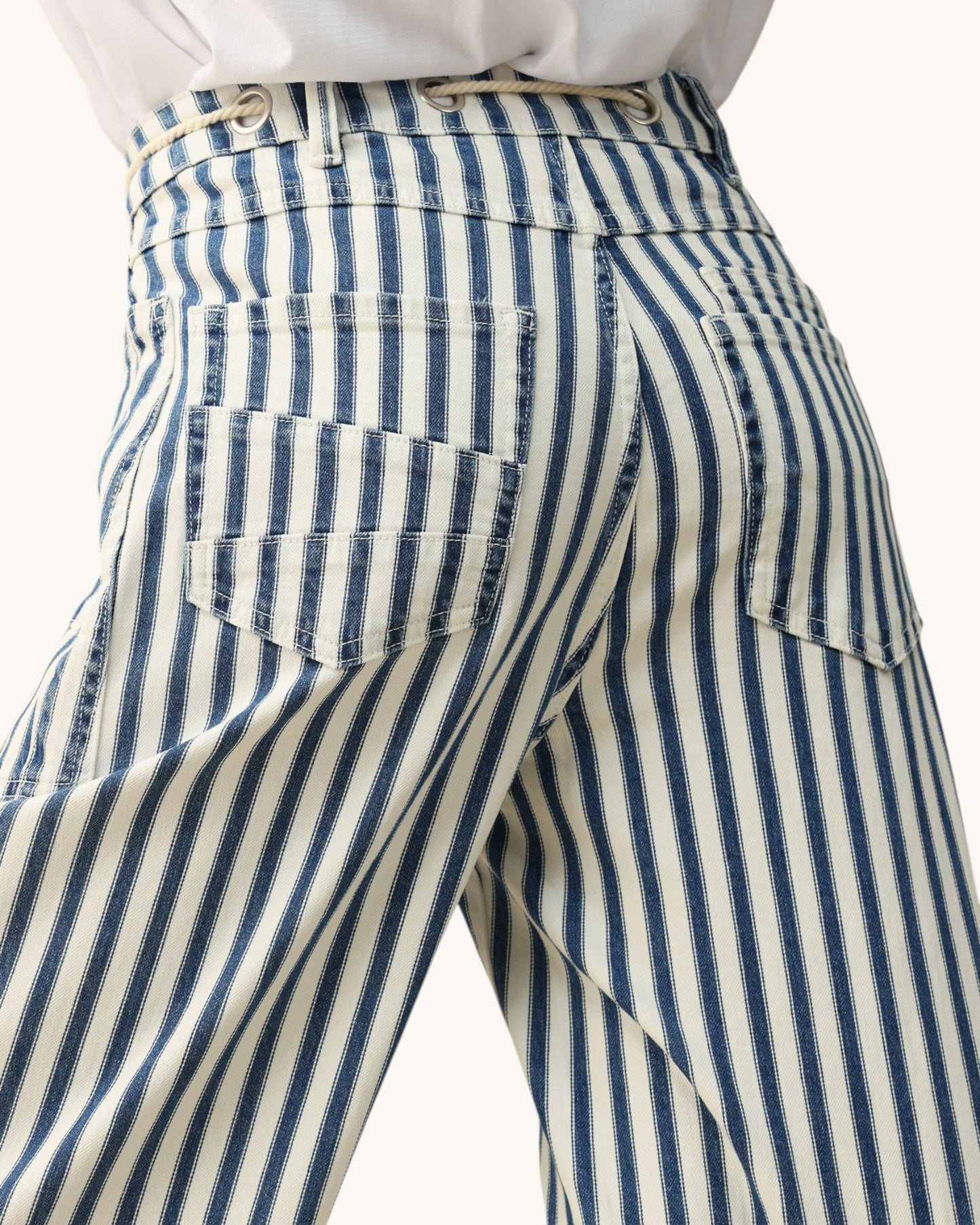 Close-up of the back waistband and pockets of striped trousers with vertical navy and cream stripes, worn with a white top against a light neutral background.