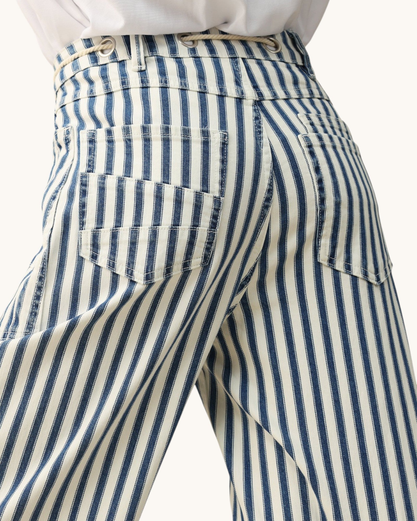 Close-up of the back waistband and pockets of striped trousers with vertical navy and cream stripes, worn with a white top against a light neutral background.