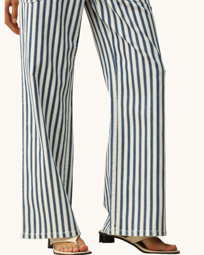 A close-up of a person wearing wide-leg pants with vertical blue and off-white stripes, paired with beige slide sandals against a plain light background.