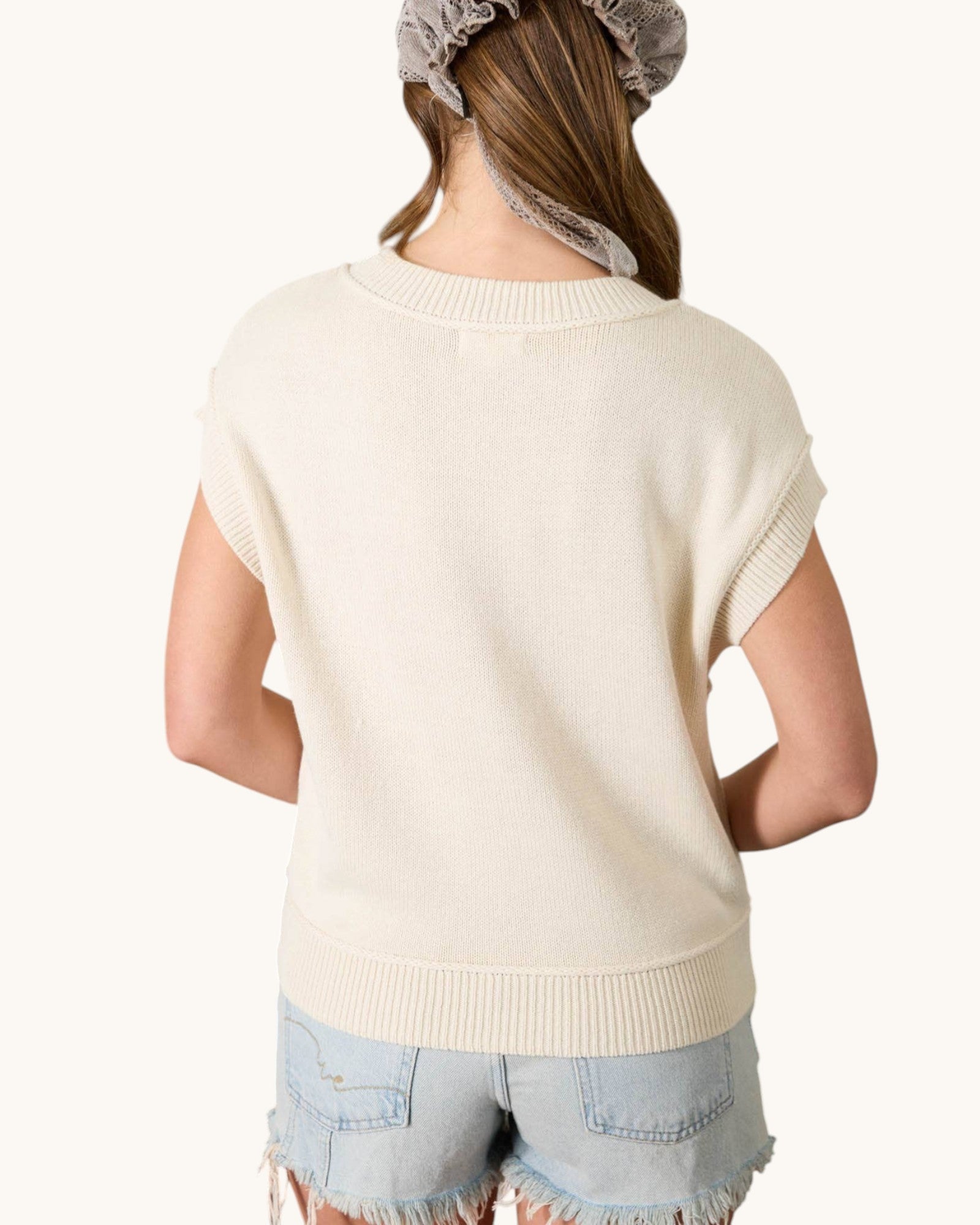 Person wearing a cream-colored sweater cardigan over a light blue top with denim shorts on a white background. 