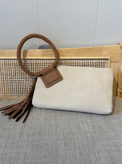 A cream-colored faux suede clutch bag with a brown braided top handle, brown leather strap, and matching tassel, displayed on a gray textured surface in front of a wooden cane panel.