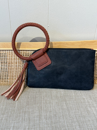 A  black suede zip pouch bag with a brown braided rope handle, a brown leather tab, and a matching brown tassel, displayed on a woven cane panel and textured beige fabric.