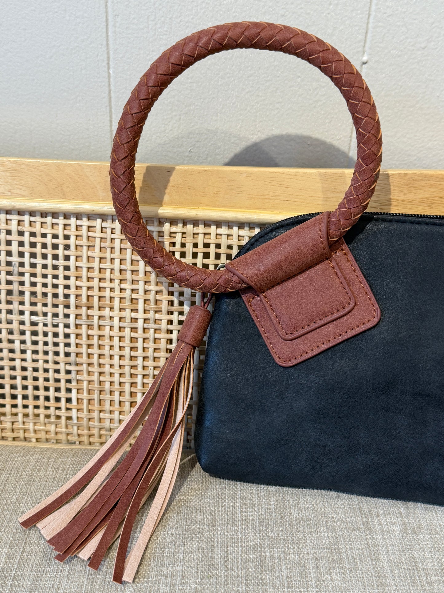 A close-up of a handbag featuring a black suede/leather body, a brown braided leather top handle, and a matching brown leather tassel with light tan fringe, set against a woven cane panel background.